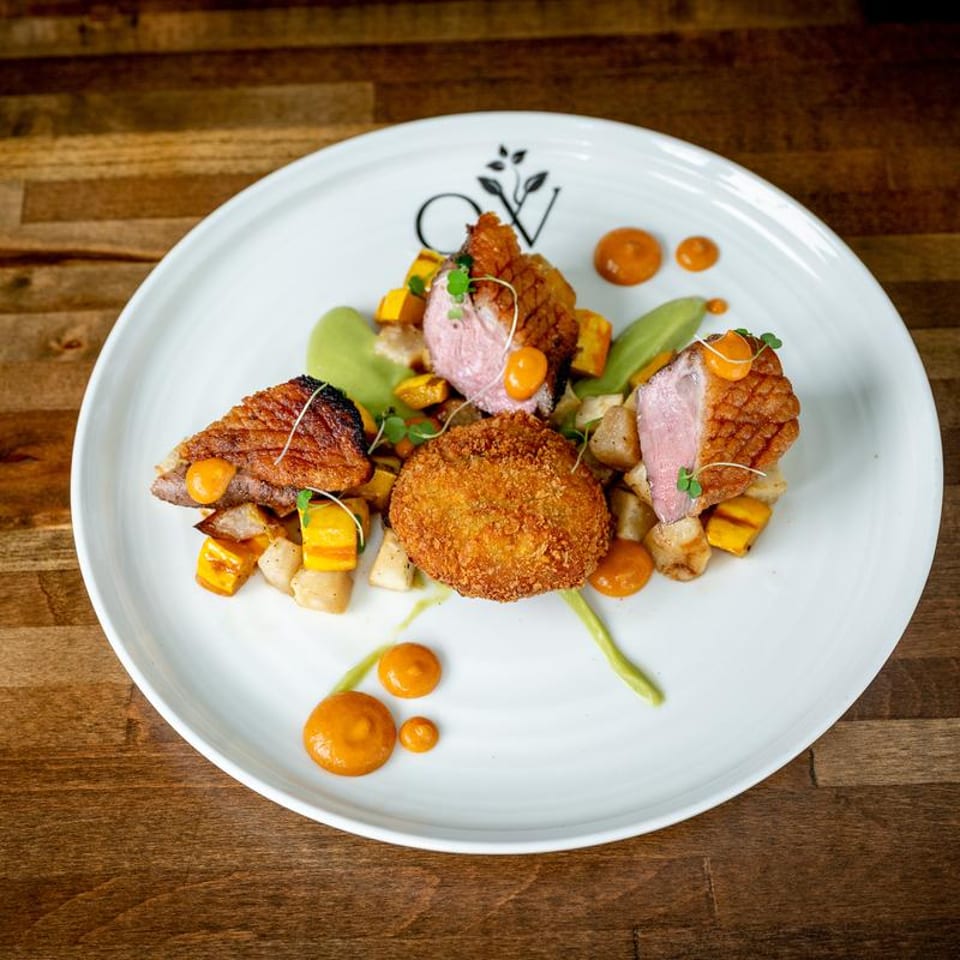 Seared duck breast with crispy croquette, roasted vegetables, green puree, and orange sauce on a white plate.
