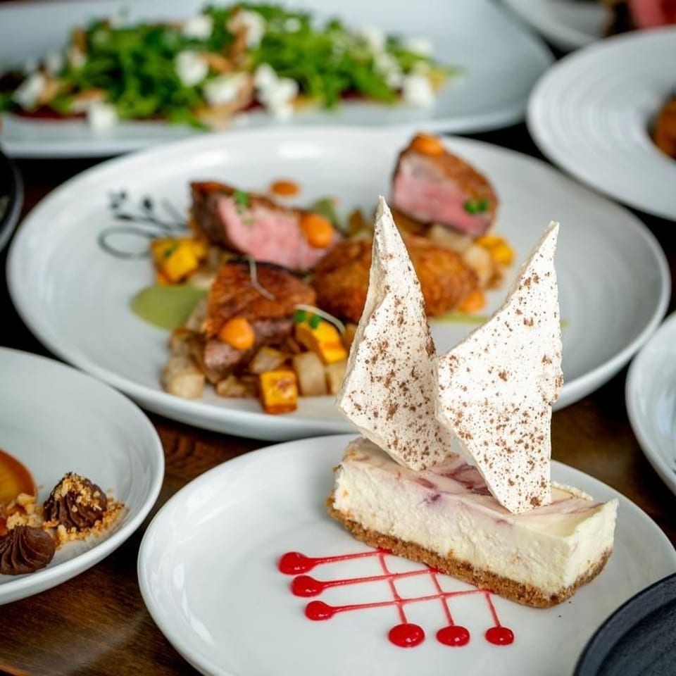 A slice of cheesecake with meringue shards and raspberry drizzle, with steak and salad in the background.