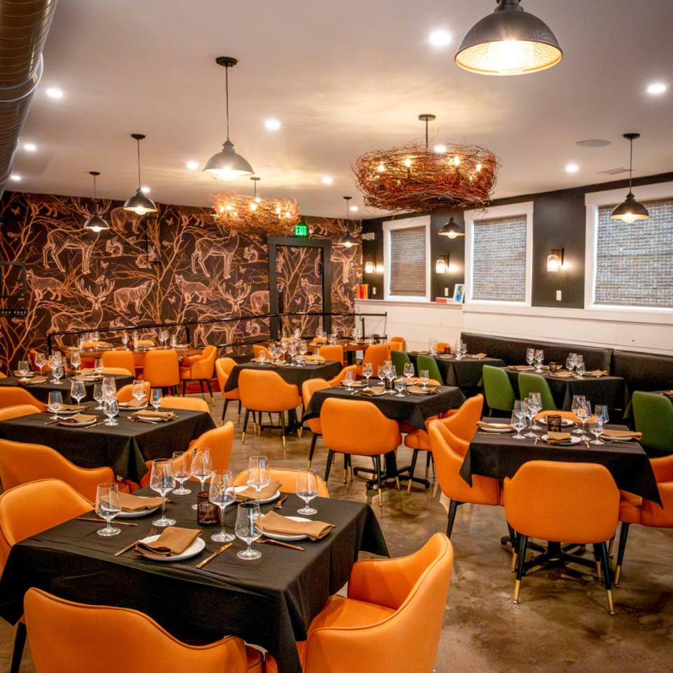 Restaurant dining room with orange chairs, black tables, and deer-patterned wallpaper under warm lighting.